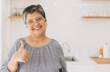 Señora mayor sonríe y muestra un pulgar arriba en medio de una cocina iluminada.