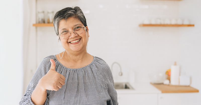 Señora mayor sonríe y muestra un pulgar arriba en medio de una cocina iluminada.