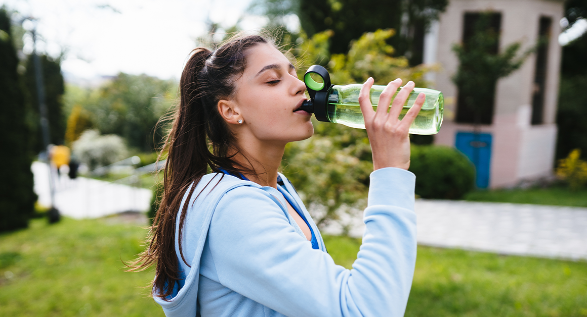 Una mujer con ropa deportiva toma agua al aire libre, representando una correcta hidratación en temporada de calor.