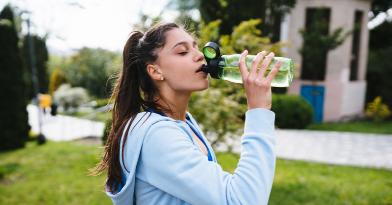 Una mujer con ropa deportiva toma agua al aire libre, representando una correcta hidratación en temporada de calor.
