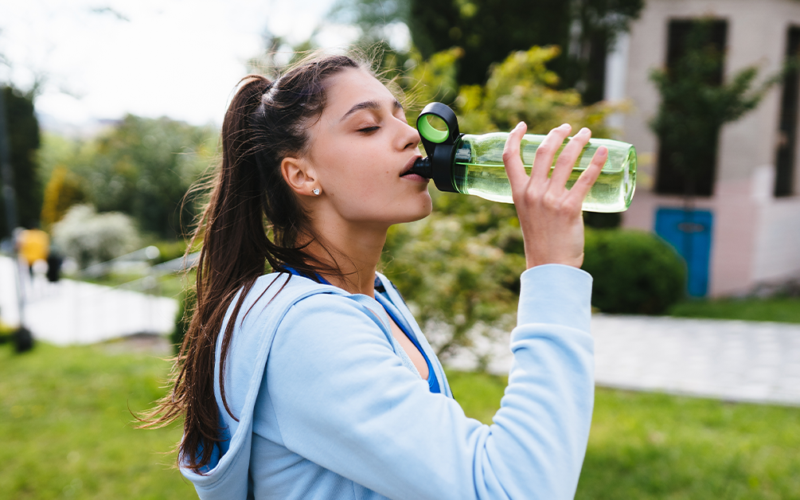 Una mujer con ropa deportiva toma agua al aire libre, representando una correcta hidratación en temporada de calor.