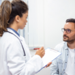 Un profesional de la salud en bata blanca sostiene un formato y conversa con un paciente sentado.