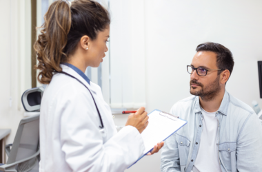 Un profesional de la salud en bata blanca sostiene un formato y conversa con un paciente sentado.