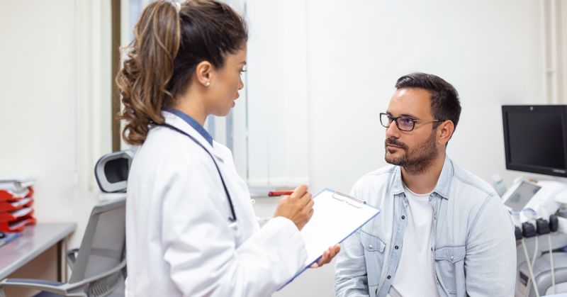 Un profesional de la salud en bata blanca sostiene un formato y conversa con un paciente sentado.