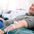 Un hombre sonriendo mientras dona sangre, reclinado en una silla. Un tubo médico está conectado a su brazo, representando el Día Mundial del Donante de Sangre.