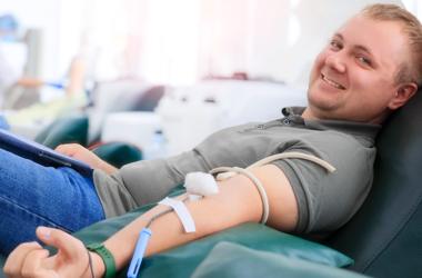 Un hombre sonriendo mientras dona sangre, reclinado en una silla. Un tubo médico está conectado a su brazo, representando el Día Mundial del Donante de Sangre.