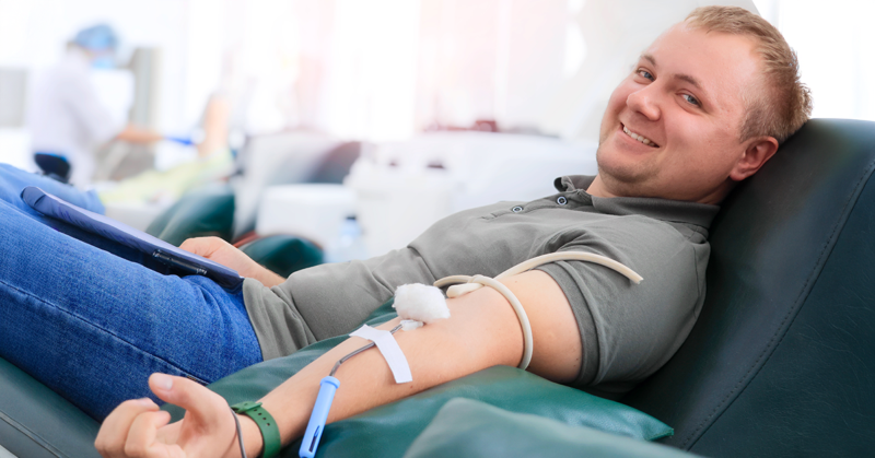 Un hombre sonriendo mientras dona sangre, reclinado en una silla. Un tubo médico está conectado a su brazo, representando el Día Mundial del Donante de Sangre.
