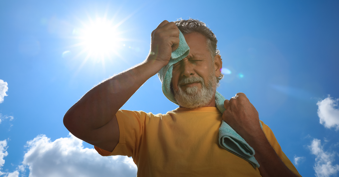 Un hombre mayor limpia el sudor de su cara con una toalla mientras está parado al aire libre, bajo el sol.