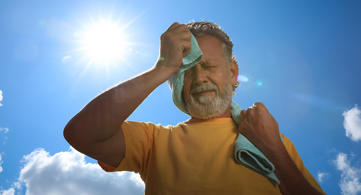 Un hombre mayor limpia el sudor de su cara con una toalla mientras está parado al aire libre, bajo el sol.
