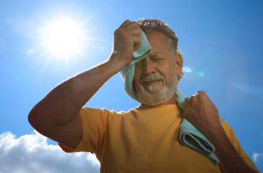 Un hombre mayor limpia el sudor de su cara con una toalla mientras está parado al aire libre, bajo el sol.