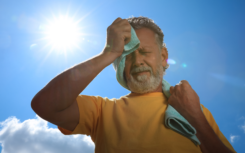 Un hombre mayor limpia el sudor de su cara con una toalla mientras está parado al aire libre, bajo el sol.