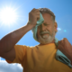 Un hombre mayor limpia el sudor de su cara con una toalla mientras está parado al aire libre, bajo el sol.