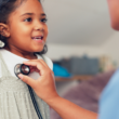 Una niña pequeña sonríe a un médico de la salud que está escuchando los latidos de su corazón con un estetoscopio.
