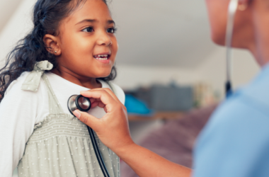 Una niña pequeña sonríe a un médico de la salud que está escuchando los latidos de su corazón con un estetoscopio.