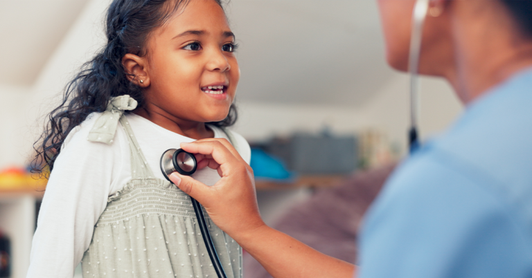 Una niña pequeña sonríe a un médico de la salud que está escuchando los latidos de su corazón con un estetoscopio.