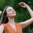 Una mujer parada al aire libre con los ojos cerrados, sonríe y alza sus brazos disfrutando de un momento de paz, representando autocuidado.