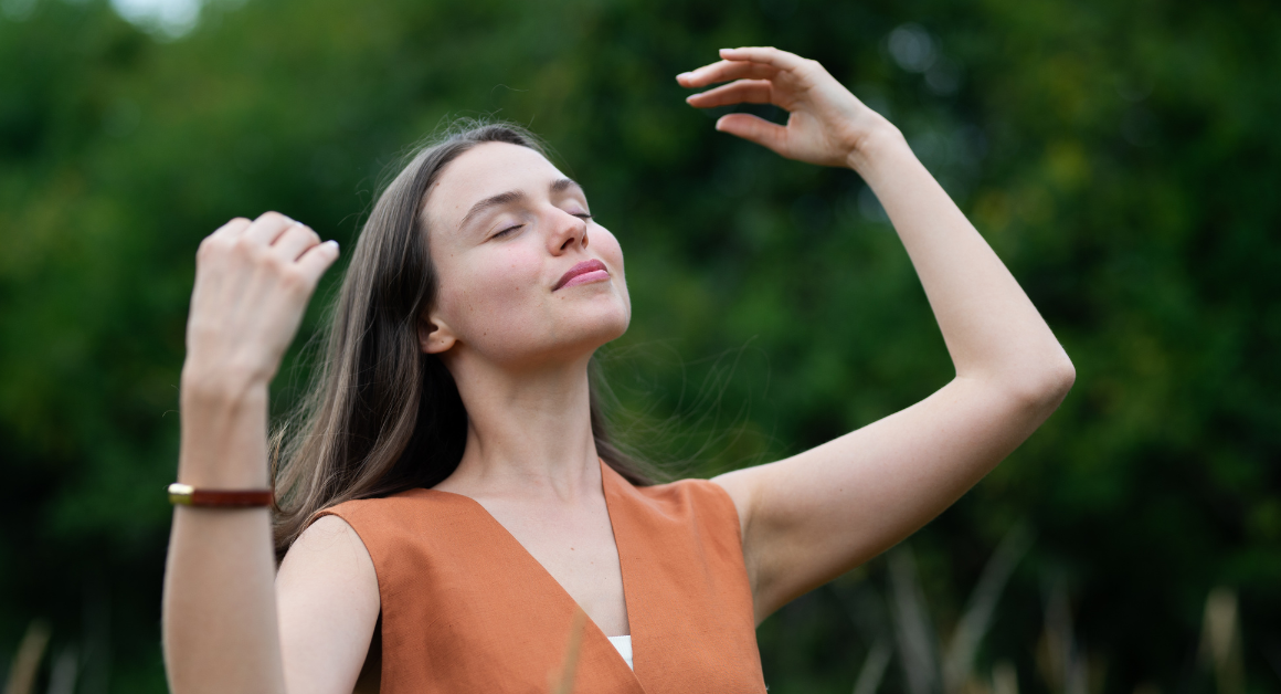 Una mujer parada al aire libre con los ojos cerrados, sonríe y alza sus brazos disfrutando de un momento de paz, representando autocuidado.