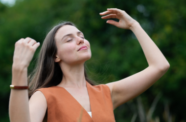 Una mujer parada al aire libre con los ojos cerrados, sonríe y alza sus brazos disfrutando de un momento de paz, representando autocuidado.