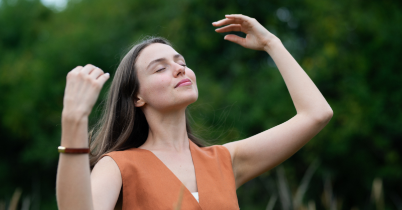 Una mujer parada al aire libre con los ojos cerrados, sonríe y alza sus brazos disfrutando de un momento de paz, representando autocuidado.