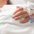 Una persona acostada en una cama de hospital descansa su mano sobre una sábana blanca, tiene una vía intravenosa sujeta con adhesivos, representando el Día Nacional de las Enfermedades Crónicas.