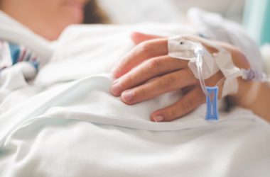 Una persona acostada en una cama de hospital descansa su mano sobre una sábana blanca, tiene una vía intravenosa sujeta con adhesivos, representando el Día Nacional de las Enfermedades Crónicas.