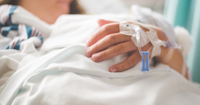 Una persona acostada en una cama de hospital descansa su mano sobre una sábana blanca, tiene una vía intravenosa sujeta con adhesivos, representando el Día Nacional de las Enfermedades Crónicas.