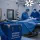 Un equipo de cirujanos llevando a cabo un trasplante de órganos mientras un miembro del equipo lleva una hielera en donde se lee "Human Organ for Donation," o en español "Órgano humano para donación."