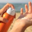 Manos aplicando protector solar de un bote naranja. En el fondo se ve una playa con arena y el mar, representando concientización sobre el cuidado de la piel.