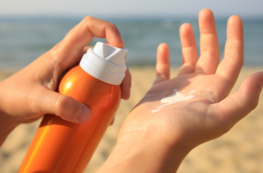 Manos aplicando protector solar de un bote naranja. En el fondo se ve una playa con arena y el mar, representando concientización sobre el cuidado de la piel.