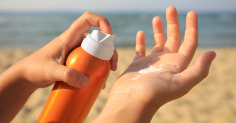 Manos aplicando protector solar de un bote naranja. En el fondo se ve una playa con arena y el mar, representando concientización sobre el cuidado de la piel.