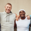 Cuatro hombres sonríen y posan con los brazos entrelazados frente a una pared beige. Visten ropa deportiva casual y transmiten compañerismo, diversidad y alegría, representando el Mes de la Salud del Hombre.
