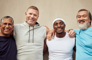 Cuatro hombres sonríen y posan con los brazos entrelazados frente a una pared beige. Visten ropa deportiva casual y transmiten compañerismo, diversidad y alegría, representando el Mes de la Salud del Hombre.