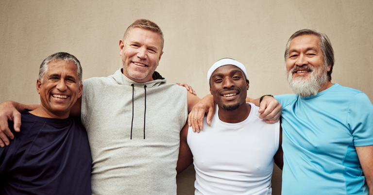 Cuatro hombres sonríen y posan con los brazos entrelazados frente a una pared beige. Visten ropa deportiva casual y transmiten compañerismo, diversidad y alegría, representando el Mes de la Salud del Hombre.