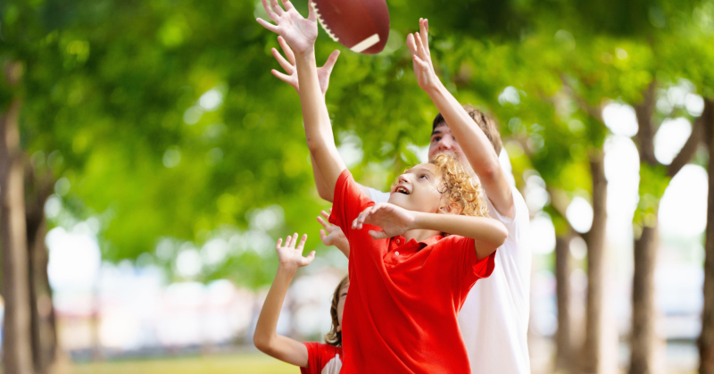 Escena energética de niños jugando a atrapar un balón de fútbol, rodeados de árboles en un parque.