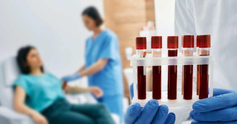 Un profesional de la salud sostiene una bandeja con pruebas de sangre. En el fondo se puede ver a una enfermera preparando a una paciente para sacarle sangre.