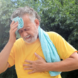 Hombre mayor en playera amarilla se limpia su sudor con una toalla. Aparece estresado, con dolor en el pecho.