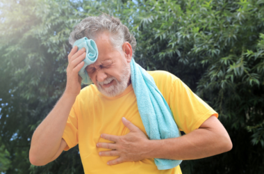 Hombre mayor en playera amarilla se limpia su sudor con una toalla. Aparece estresado, con dolor en el pecho.