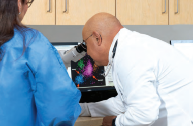 El Dr. Jesús Gómez, hematólogo y oncólogo médico certificado por el consejo de especialidad, examina imágenes de datos multicolores mediante un microscopio conectado a una pantalla de computadora.