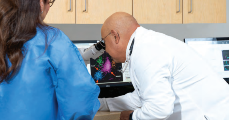 El Dr. Jesús Gómez, hematólogo y oncólogo médico certificado por el consejo de especialidad, examina imágenes de datos multicolores mediante un microscopio conectado a una pantalla de computadora.