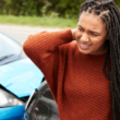 Mujer parada frente a un auto con el frente abollado, se frota el cuello con dolor, representando concientización sobre el trauma.