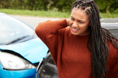 Mujer parada frente a un auto con el frente abollado, se frota el cuello con dolor, representando concientización sobre el trauma.