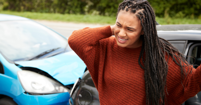 Mujer parada frente a un auto con el frente abollado, se frota el cuello con dolor, representando concientización sobre el trauma.