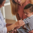 Un profesional de la salud se prepara para vacunar a una niña pequeña, mientras su madre la sostiene.
