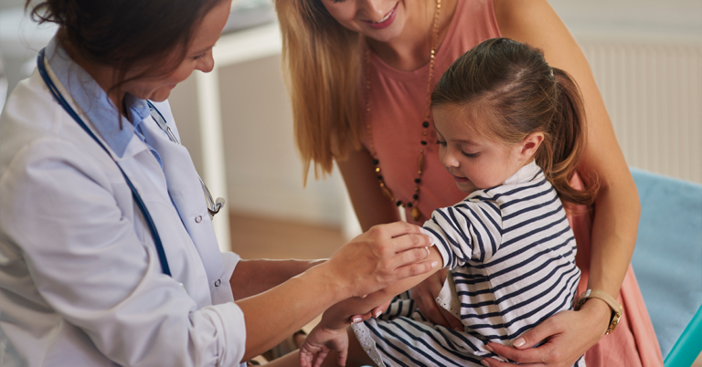 Un profesional de la salud se prepara para vacunar a una niña pequeña, mientras su madre la sostiene.