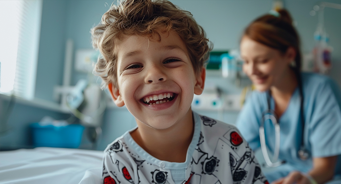 Niño sonriendo en una cama de hospital con una enfermera detrás.