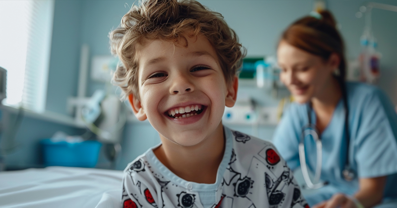 Niño sonriendo en una cama de hospital con una enfermera detrás.