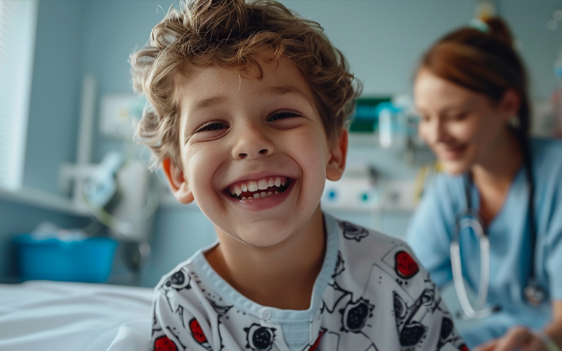 Niño sonriendo en una cama de hospital con una enfermera detrás.