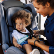 Mujer sonríe mientras abrocha el arnés del asiento de auto de un niño pequeño en el asiento trasero de un vehículo