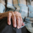 Mano de una persona mayor apoyada sobre su rodilla, con camisa de cuadros y pantalón gris