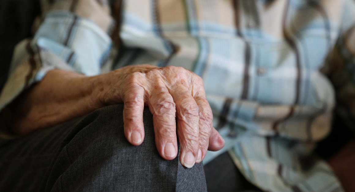 Mano de una persona mayor apoyada sobre su rodilla, con camisa de cuadros y pantalón gris
